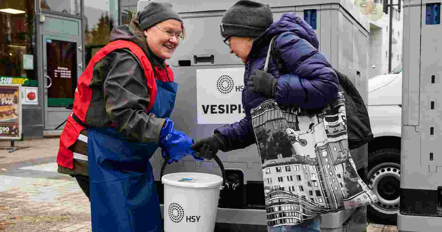 Hymyilevä henkilö Punaisen Ristin vapaaehtoisliiveissä ojentaa kannellista ämpäriä naiselle, jonka vedenjakelu on katkennut. Punainen Risti suorittaa vedenjakelua kotimaan kriisitilanteissa.